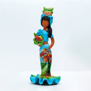 May include: A handcrafted figurine of a woman in a blue outfit, carrying a basket of produce and a stack of food on her head. The skirt features a painted landscape with a tree. The figurine has dark hair and skin, and a ruffled base.