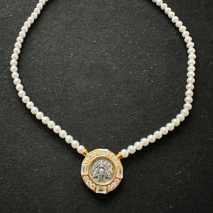 May include: A white pearl necklace with a gold pendant. The pendant features a silver coin with a bee design, surrounded by clear rectangular and round crystals. The necklace is displayed on a black surface.