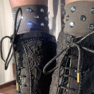 Peut inclure: Bottes noires à motifs de dentelle avec des lacets noirs et des embouts dorés. Les bottes sont montantes et sont associées à des chaussettes grises ornées de strass étincelants. Les bottes sont ornées de motifs floraux complexes.