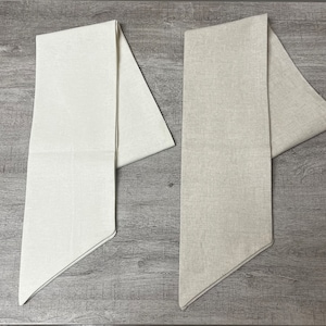 May include: Two beige linen table runners, one folded in half and the other folded in thirds, are laid out on a wood surface.