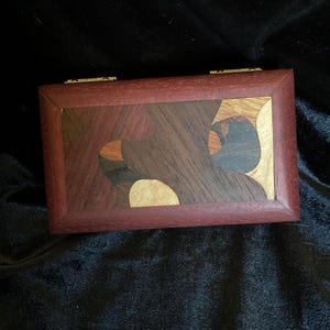 May include: A small wooden box with a hinged lid. The box is made of various types of wood, including dark brown, reddish brown, and light brown. The lid is decorated with a geometric pattern of different colored woods.