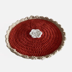 May include: A round crocheted placemat with a scalloped edge in orange and beige with a white crocheted flower in the center.