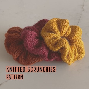 Può includere: Tre scrunchies di lana in diversi colori: giallo senape, bordeaux e arancione bruciato. Gli scrunchies sono impilati uno sopra l'altro.  KNITTED SCRUNCHIES PATTERN è scritto in testo nero su sfondo rosa.