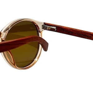 May include: Close-up of round sunglasses with clear frames and dark lenses. The temples are made of dark wood, with the text "Polarized Rosewood" visible. The sunglasses are set against a white background.