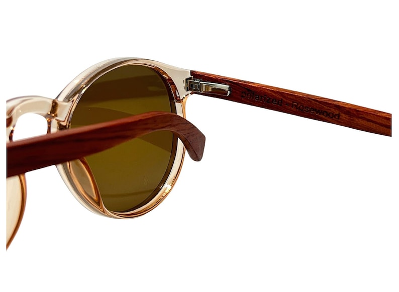 May include: Close-up of round sunglasses with clear frames and dark lenses. The temples are made of dark wood, with the text "Polarized Rosewood" visible. The sunglasses are set against a white background.