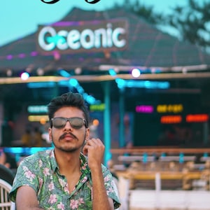 May include: A person wearing sunglasses and a floral shirt sits in a chair. The word "Yoguni" is written in cursive at the top of the image. The sign behind the person says "Ceeonic". The background is a beach setting.