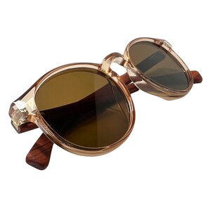 May include: Round sunglasses with brown lenses and a light brown frame. The temples are made of dark wood. The frame has a decorative crystal-like accent at the hinge.