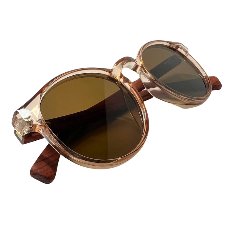 May include: Round sunglasses with brown lenses and a light brown frame. The temples are made of dark wood. The frame has a decorative crystal-like accent at the hinge.