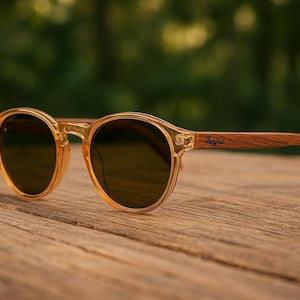 May include: Round sunglasses with a translucent amber frame and dark lenses. The temples are made of wood and feature a brand name. The sunglasses rest on a wooden surface, with a blurred green background.