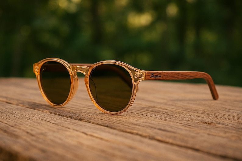 May include: Round sunglasses with a translucent amber frame and dark lenses. The temples are made of wood and feature a brand name. The sunglasses rest on a wooden surface, with a blurred green background.