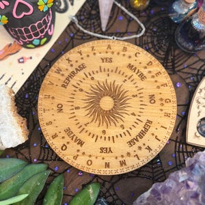 May include: A wooden Ouija board with engraved letters, numbers, and the words "YES," "NO," and "MAYBE." The board features a sunburst design in the center and is surrounded by other mystical items, including crystals and decorative bottles.