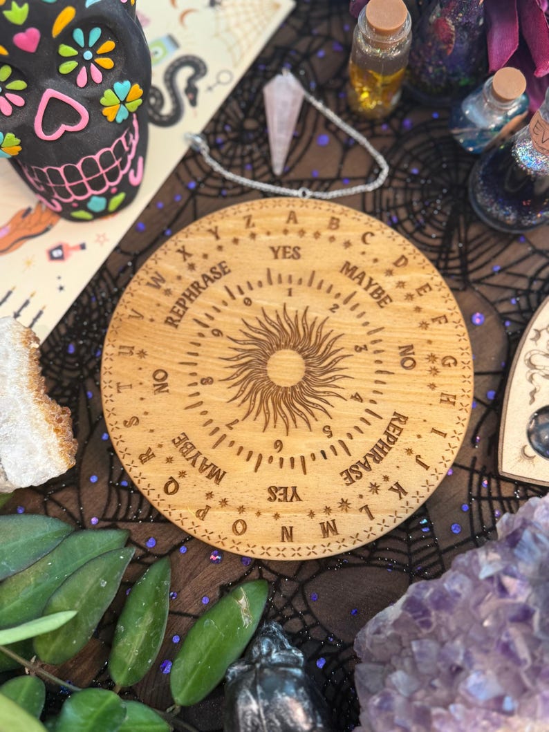 May include: A wooden Ouija board with engraved letters, numbers, and the words "YES," "NO," and "MAYBE." The board features a sunburst design in the center and is surrounded by other mystical items, including crystals and decorative bottles.