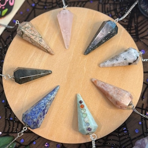 May include: A collection of eight gemstone pendulums on a round wooden surface. The pendulums are various colors, including pink, blue, and green, and are suspended from silver chains. The background features a dark fabric with a spiderweb design.