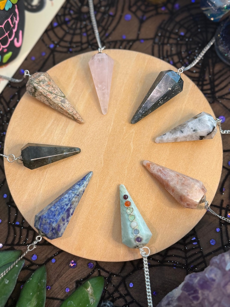 May include: A collection of eight gemstone pendulums on a round wooden surface. The pendulums are various colors, including pink, blue, and green, and are suspended from silver chains. The background features a dark fabric with a spiderweb design.