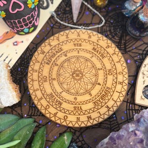 May include: A wooden divination board with engraved letters, numbers, and symbols, including an eye design. The board features the words "YES," "NO," and "MAYBE." The board is surrounded by other mystical items, including a decorative skull and crystals.