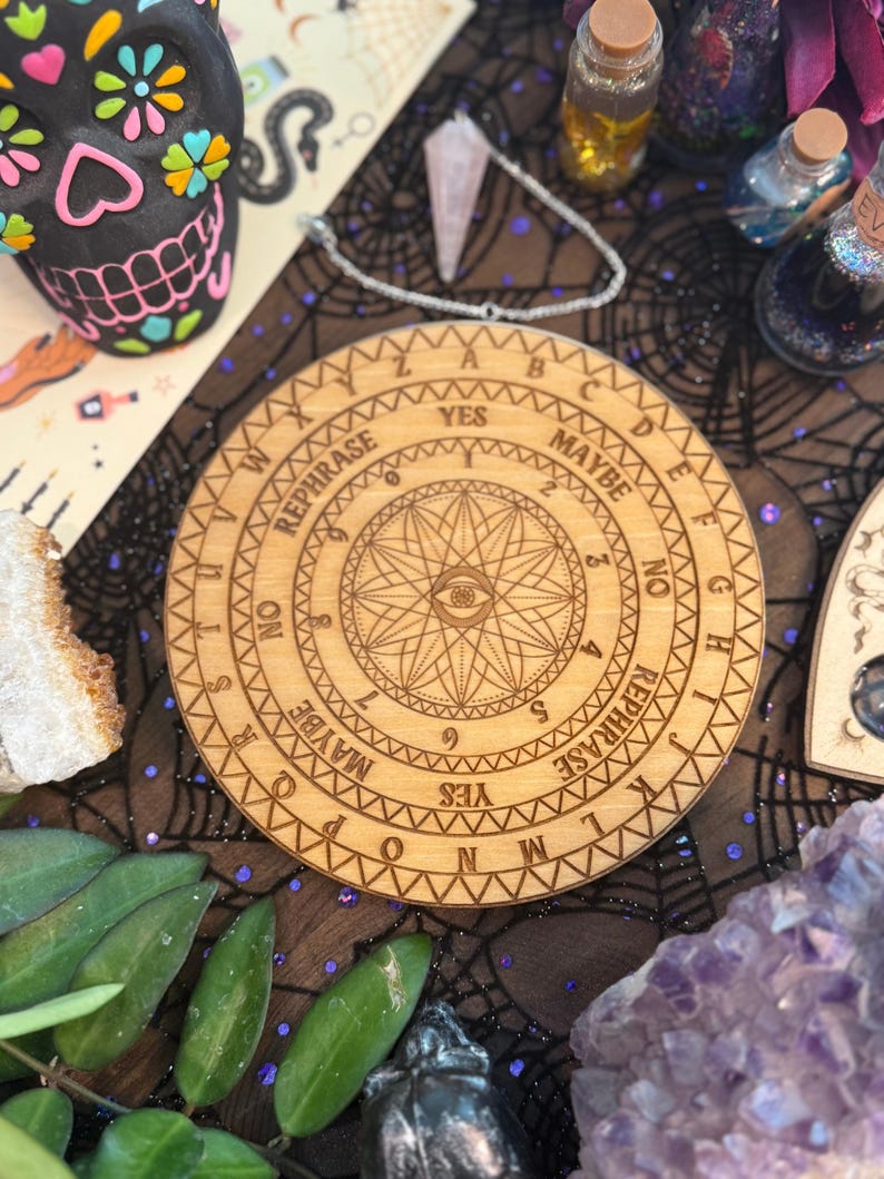 May include: A wooden divination board with engraved letters, numbers, and symbols, including an eye design. The board features the words "YES," "NO," and "MAYBE." The board is surrounded by other mystical items, including a decorative skull and crystals.