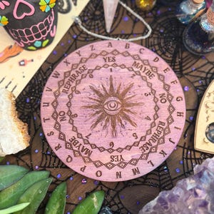 May include: A round, light purple wooden Ouija board with engraved letters, numbers, and phrases. The board features a central sunburst design with an eye. The word "YES" is prominently displayed. The board is surrounded by other mystical items.