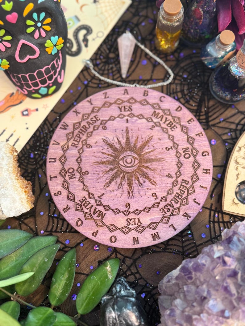 May include: A round, light purple wooden Ouija board with engraved letters, numbers, and phrases. The board features a central sunburst design with an eye. The word "YES" is prominently displayed. The board is surrounded by other mystical items.