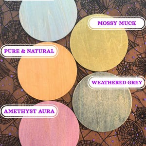 May include: Five round wooden coasters in various colors: Frozen Sky, Mossy Muck, Pure & Natural, Weathered Grey, and Amethyst Aura. Each coaster has a unique wood grain pattern. The coasters are displayed on a black and brown spiderweb-patterned fabric.