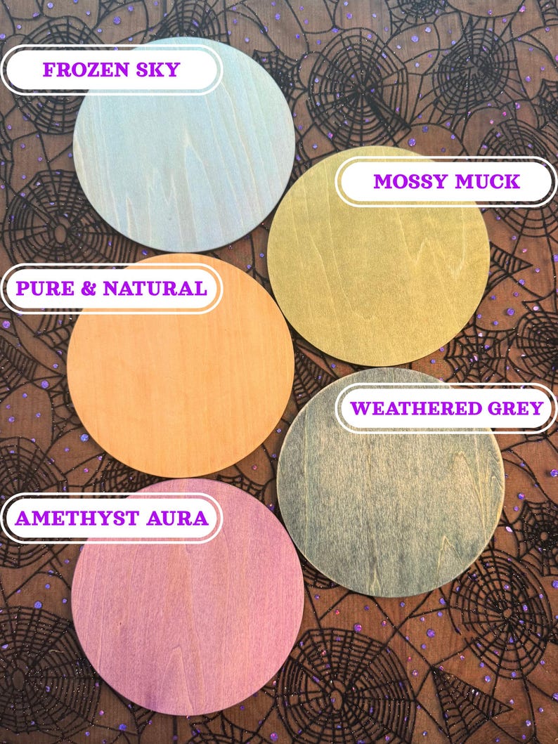 May include: Five round wooden coasters in various colors: Frozen Sky, Mossy Muck, Pure & Natural, Weathered Grey, and Amethyst Aura. Each coaster has a unique wood grain pattern. The coasters are displayed on a black and brown spiderweb-patterned fabric.