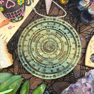 May include: A wooden planchette with engraved letters, numbers, and phrases like "YES" and "MAYBE". The planchette has a circular design with a central illustration of a crescent moon and a star. The planchette is surrounded by other mystical items.