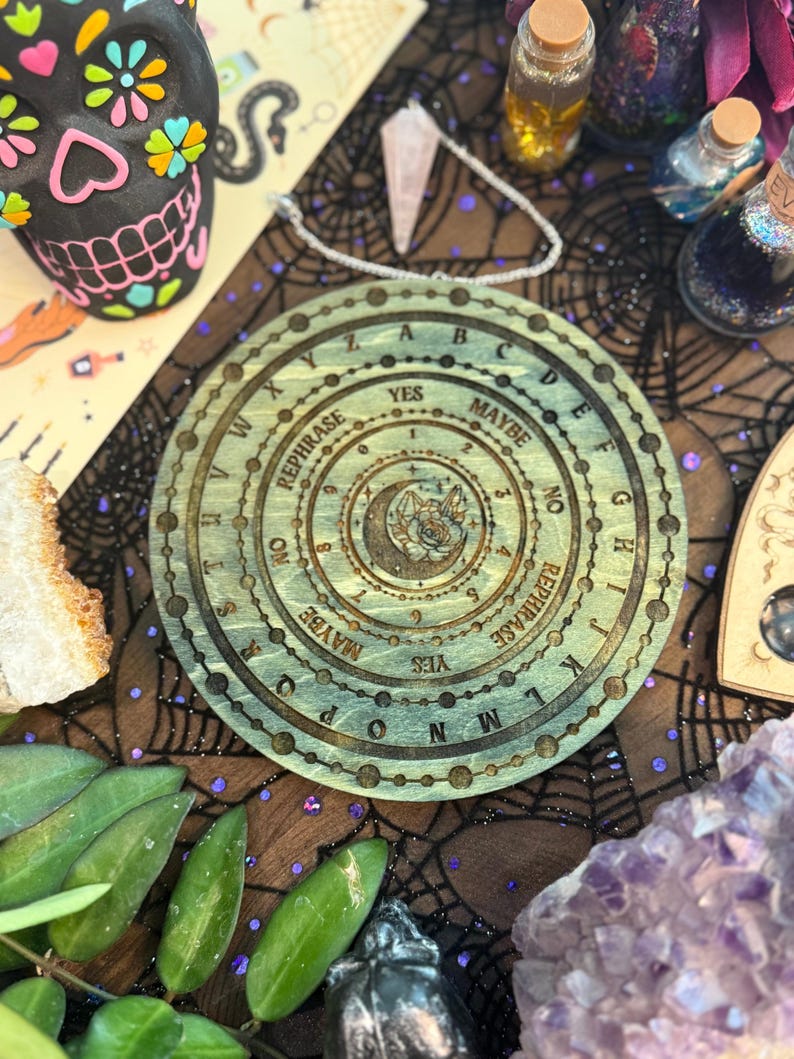 May include: A wooden planchette with engraved letters, numbers, and phrases like "YES" and "MAYBE". The planchette has a circular design with a central illustration of a crescent moon and a star. The planchette is surrounded by other mystical items.