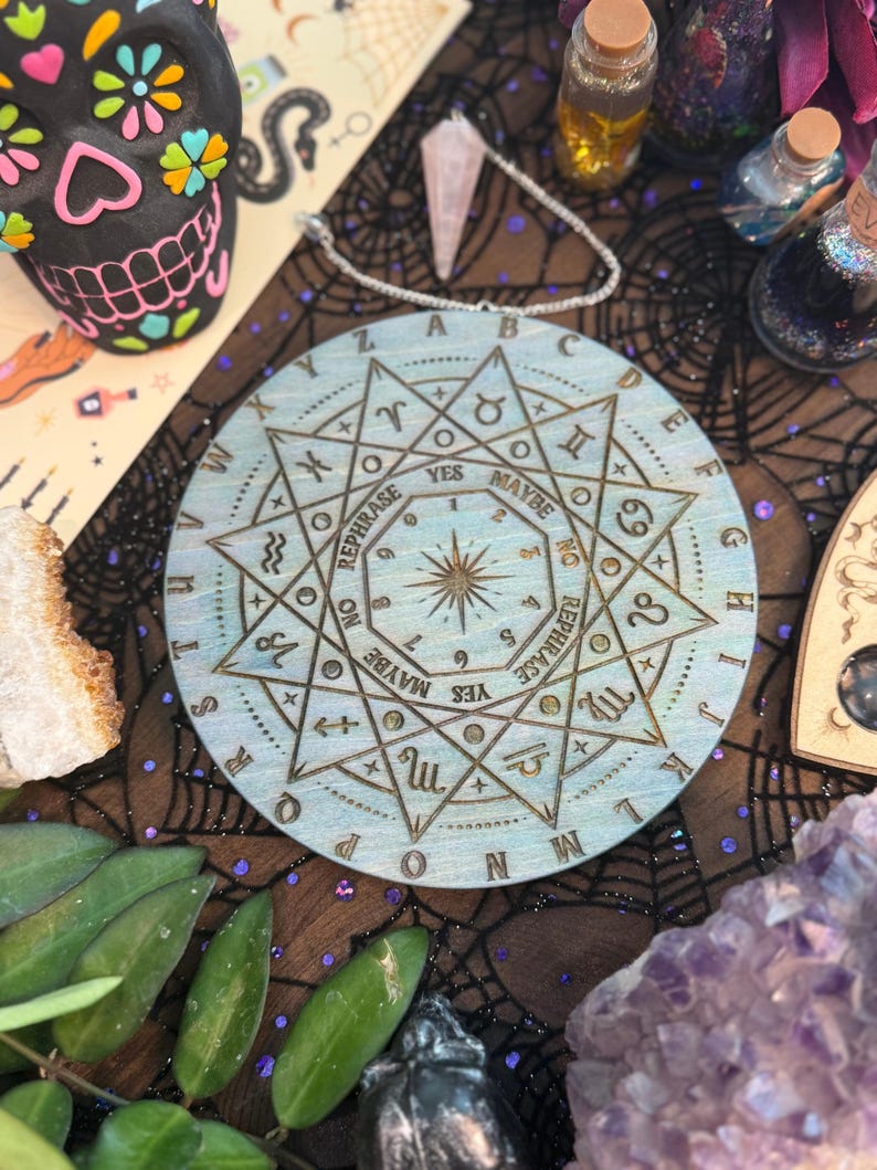 May include: A round, light blue wooden divination board with engraved symbols, letters, and the words "YES," "MAYBE," and "REPHRASE." A starburst design is in the center. A rose quartz crystal hangs above the board.
