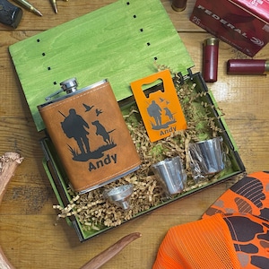 May include: A green wooden box with a brown leather flask, two shot glasses, and an orange bottle opener. The flask and opener feature a hunting-themed design with the name "Andy". Also included are shotgun shells and a hunting hat.
