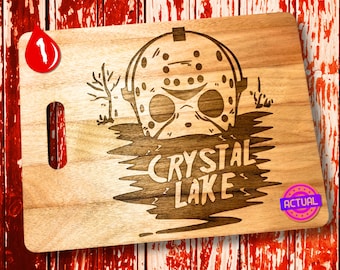 May include: A wooden chopping board with a laser-engraved design of a hockey mask and the text "Crystal Lake". The design is in brown and black on a light brown wooden surface. The board has a handle on the side.