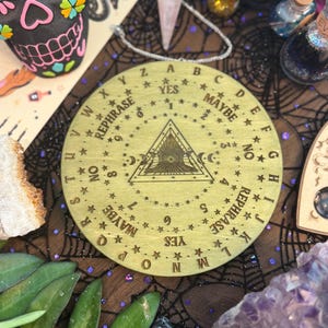 May include: A round, wooden planchette board with engraved letters, numbers, and phrases like "Yes," "No," and "Maybe." The board features a central triangle design and is surrounded by stars. The board is on a dark surface with other mystical items.