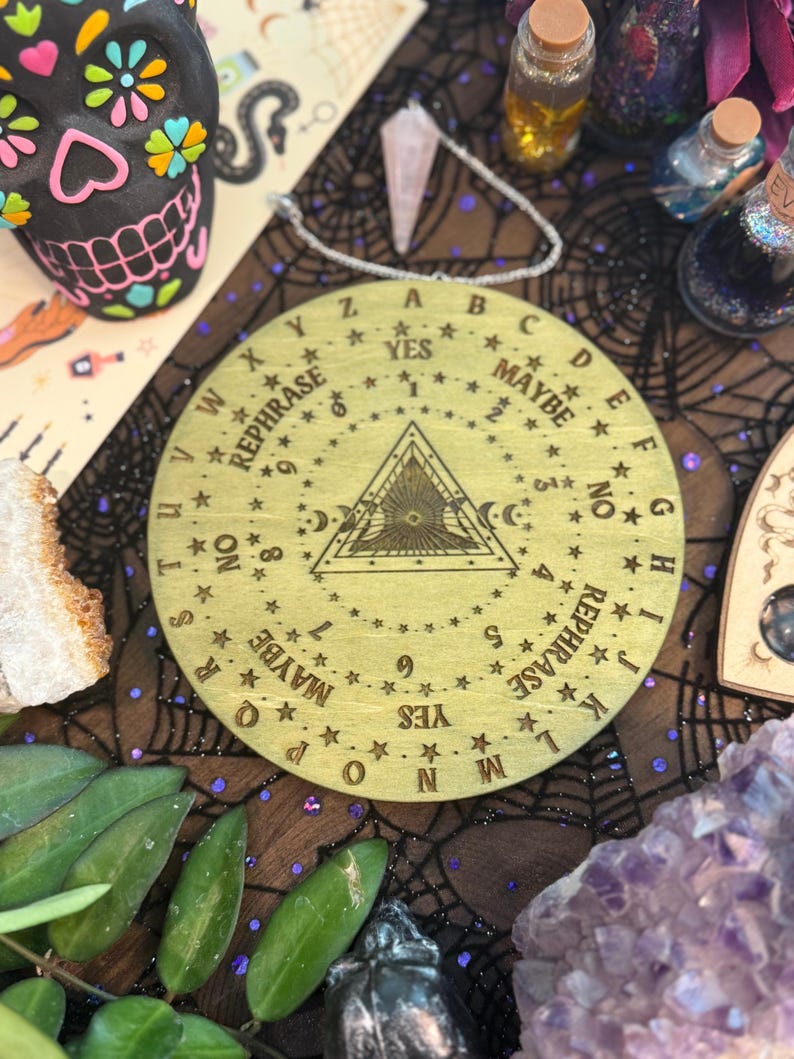 May include: A round, wooden planchette board with engraved letters, numbers, and phrases like "Yes," "No," and "Maybe." The board features a central triangle design and is surrounded by stars. The board is on a dark surface with other mystical items.