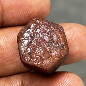 Könnte beinhalten: Ein roher, rotbrauner Edelstein mit sechseckiger Form. Die Oberfläche hat ein strukturiertes Aussehen mit Farb- und Tonvariationen. Der Edelstein wird in einer Hand gehalten und zeigt seinen natürlichen, unpolierten Zustand.