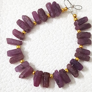 May include: A necklace featuring raw, dark pink ruby gemstone beads. The beads are strung together with gold-colored spacers and a silver-colored clasp. The necklace is displayed on a white surface.