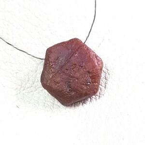 以下が含まれることがあります： 薄い黒いワイヤーに吊るされた、六角形の未加工ルビーの宝石。 ルビーは、表面の質感とファセットが目立つ、濃い赤茶色の色合いをしています。 宝石は白い背景に配置されています。