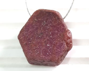 May include: A rough, uncut ruby gemstone with a deep red hue. The natural stone is irregularly shaped with a textured surface and is suspended by a thin, silver-colored wire. The background is white.