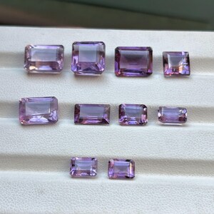 May include: A collection of rectangular-cut amethyst gemstones in various sizes. The gems exhibit a spectrum of purple shades, from pale lavender to rich violet, with a polished, reflective finish. The gemstones are displayed on a white surface.
