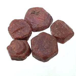 Puede incluir: Cinco piedras preciosas de rubí en bruto sin cortar. Las piedras son de un color marrón rojizo intenso con formas irregulares y superficies texturizadas. Las piedras preciosas se muestran sobre un fondo blanco.
