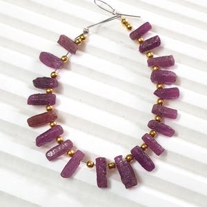 May include: A necklace featuring raw, irregular-shaped, deep pink gemstones strung with gold-colored beads. The gemstones are a rich, purplish-pink hue, and the necklace is displayed against a white background.