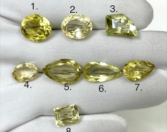 May include: An assortment of eight yellow gemstones in various shapes, including oval, pear, and emerald cuts. The gems are displayed on a white surface, showcasing their clarity and color.
