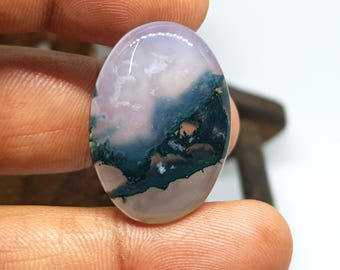 May include: Oval-shaped gemstone with a polished surface. The stone features a blend of colors, including pale lavender, white, and deep teal, with hints of orange. The design resembles a landscape or abstract art. The gemstone is held between fingers.