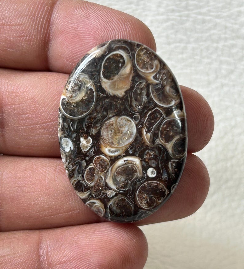 K&ouml;nnte beinhalten: Ovaler Cabochon-Edelstein mit braunem und wei&szlig;em Muster. Der Stein hat ein wirbelndes, organisches Design mit vielen kleinen Kreisen und Ovalen.