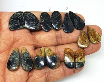 May include: Assortment of seven pairs of polished gemstones in various shapes. The stones display a range of colors, including black, brown, and tan, with unique veining patterns. The gemstones are held in a hand against a white background.