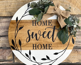 May include: A round wooden sign with a white and brown striped background. The sign has a black wreath with leaves and the words "Home sweet home" written in black lettering. The sign is decorated with a burlap bow and greenery.