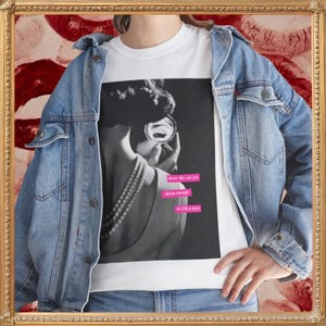 May include: A white t-shirt featuring a black and white graphic of a person applying makeup with a mirror, wearing a pearl necklace. The graphic includes pink rectangular text boxes with the words: draw the cat eye, sharp enough, to kill a man.