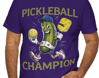 May include: A purple t-shirt with the text "PICKLEBALL CHAMPION" in gold. The design shows a cartoon pickle wearing a cap, holding a pickleball and paddle. The pickle is running and smiling, a fun design.