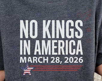May include: Dark gray t-shirt with the text "NO KINGS IN AMERICA" in large white letters. Below the text is "MARCH 28, 2026" and a graphic of a star and stripes. The shirt has a crew neck.