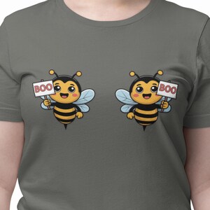 Puede incluir: Camiseta gris con dos abejas de dibujos animados, cada una sosteniendo un cartel que dice "BOO". Las abejas son negras y amarillas con alas azules y caras sonrientes. La camiseta es de color gris liso.