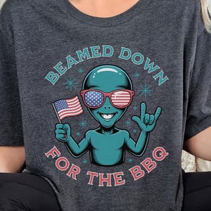 Puede incluir: Camiseta gris oscuro con un extraterrestre verde azulado que lleva gafas de sol rojas, blancas y azules. El extraterrestre sostiene una bandera estadounidense y hace un gesto de rock. El texto dice "BEAMED DOWN FOR THE BBQ" en un diseño circular.