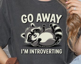 Puede incluir: Camiseta gris oscuro con el texto "GO AWAY" sobre una ilustración de mapache de dibujos animados. Debajo del mapache está el texto "I'M INTROVERTING". El mapache es en tonos grises y blancos.