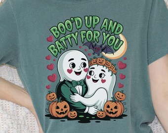 以下が含まれることがあります： ハロウィーンをテーマにしたグラフィックが特徴のティール色のTシャツ。デザインには、抱き合う2匹の幽霊、カボチャ、ハート、コウモリ、そして緑色の「BOO'D UP AND BATTY FOR YOU」という文字が含まれています。幽霊はバラ色の頬をしており、お祝いの要素に囲まれています。